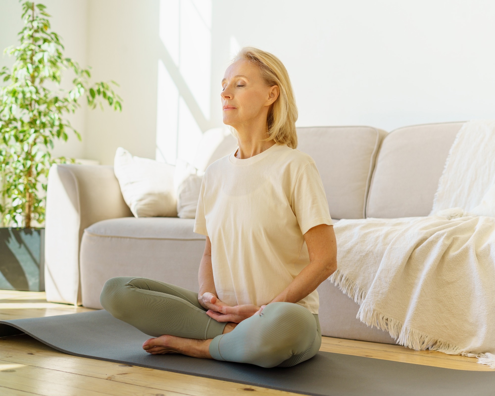 Peaceful senior woman in lotus position meditation with closed eyes at home while sitting on yoga mat on floor, full length.