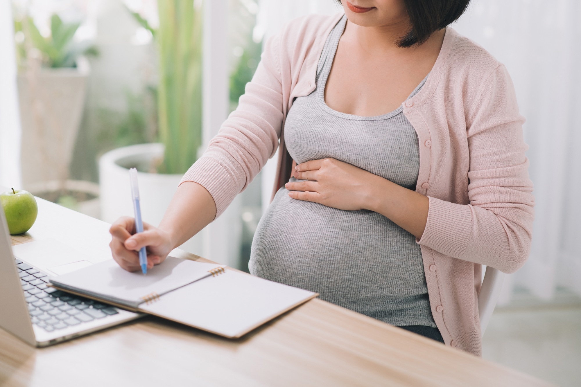 Closeup of a pregnant woman writing notes and using a laptop