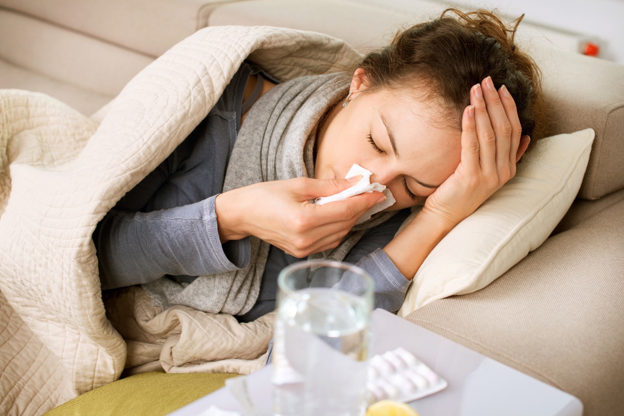 young woman with flu, lying down on sofa, clutching her had and blowing her nose. Glass of water a medication next to her
